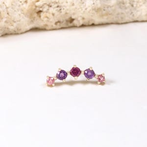 Może przedstawiać: Zakrzywiony, złoty kolczyk z rzędem kolorowych kamieni szlachetnych. Kamienie szlachetne są w odcieniach różu, fioletu i magenty. Kolczyk jest umieszczony na białym tle z teksturowanym, jasnym kamieniem.