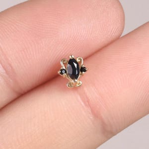 Puede incluir: Un pequeño pendiente dorado con una piedra negra central de forma ovalada y dos piedras negras más pequeñas a cada lado. El pendiente tiene un diseño decorativo y ornamentado. La joya se muestra sobre un fondo neutro.