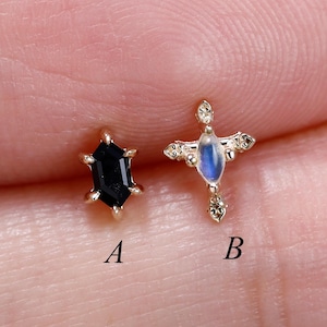 May include: Two gold stud earrings labeled A and B. Earring A features a black hexagonal gemstone in a prong setting. Earring B is a gold cross-shaped stud with a central marquise-cut iridescent moonstone and small clear accent stones.