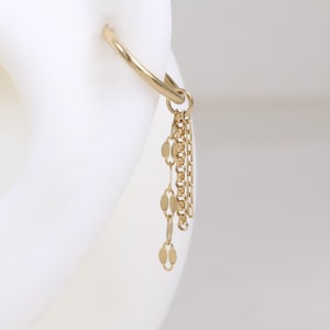 May include: A gold-colored hoop earring with a dangling chain of small, oval-shaped charms. The earring is shown on a white ear model, highlighting its design and details. The earring is a piece of jewelry.
