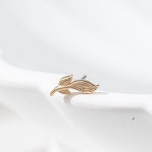 May include: A gold-colored leaf-shaped earring stud. The earring is small and delicate, with detailed veining on the leaves. The earring is set against a white background, highlighting its intricate design. The earring is a piece of jewelry.