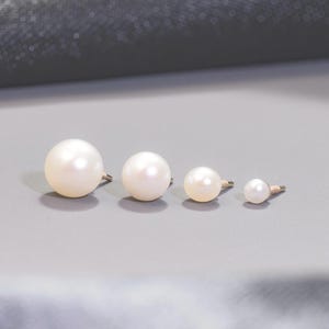 May include: Four round white pearl stud earrings arranged in a horizontal row from largest to smallest. Each pearl features a metallic post extending from the back, showcasing a smooth, lustrous surface and a classic spherical shape for each piece.