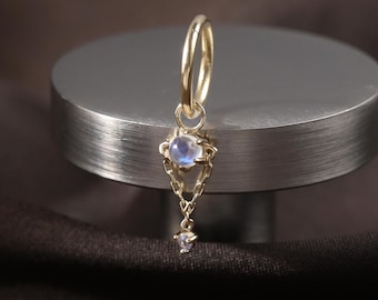May include: A single gold hoop earring with a dangling pendant. The pendant features a teardrop-shaped, light blue gemstone at the top, with a delicate chain leading to a small, clear gemstone at the bottom. The earring is displayed on a silver cylinder against a dark brown background.