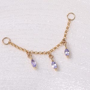 Puede incluir: Una delicada cadena dorada con tres gemas púrpuras colgantes en forma de lágrima. La cadena tiene pequeños aros en cada extremo, diseñados para ser usados como joyas. El fondo es una tela blanca texturizada.