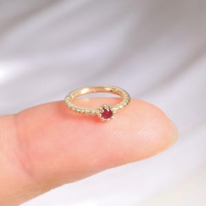 Puede incluir: Un delicado anillo de oro con una pequeña gema roja y redonda. El anillo se sostiene en un dedo, mostrando su intrincado diseño y el color vibrante de la gema. La banda del anillo tiene un patrón texturizado.