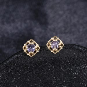 Peut inclure: Une paire de boucles d'oreilles à clous dorées. Chaque boucle d'oreille présente une pierre précieuse violet clair sertie dans un cadre carré ajouré décoratif. Les boucles d'oreilles sont présentées sur une surface sombre et texturée.