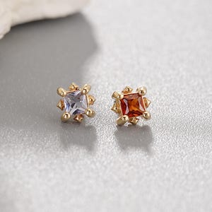 May include: A pair of gold stud earrings featuring square-cut gemstones. One earring contains a light blue stone, while the other contains a deep orange stone. Each stone is held in a four-prong setting with small decorative metal accents at each corner.