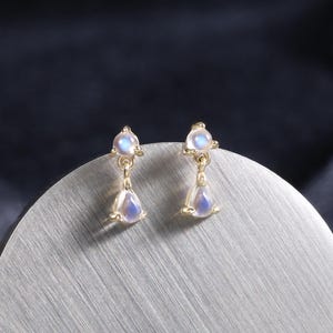 May include: A pair of gold-colored drop earrings with a small, round, light blue gemstone at the top and a teardrop-shaped gemstone hanging below. The earrings are displayed on a silver, circular surface against a dark background.