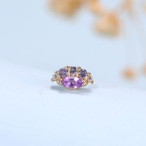 May include: A gold stud earring featuring a central marquise-cut purple gemstone surrounded by a semi-circular arrangement of smaller, clear round-cut stones. The piece is crafted with a delicate metal setting that holds the stones in place.