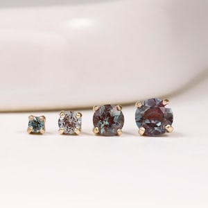 May include: Four round-cut alexandrite gemstones set in gold prongs, arranged in a horizontal line from smallest to largest. Each stone displays a colour-shifting hue ranging from deep purple and blue to teal, secured by four small gold metal prongs.