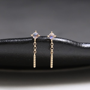 Könnte beinhalten: Ein Paar goldfarbene Ohrringe mit einem kleinen, runden, hellblauen Edelstein. Jeder Ohrring hat eine zarte Kette mit einem vertikalen Stab aus kleinen, funkelnden Akzenten. Präsentiert auf einer schwarzen, strukturierten Oberfläche.