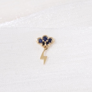 May include: A gold-colored earring featuring a cloud design with five dark blue gemstones and a dangling lightning bolt. The earring is set against a white, textured background.