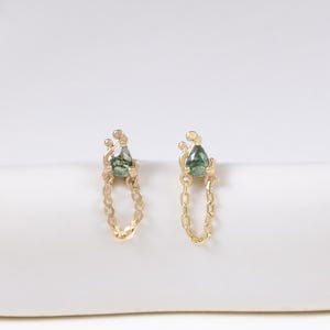May include: A pair of gold chain earrings with a small green gemstone at the top and bottom. The earrings are displayed on a white, curved surface. The gemstones have a teardrop shape.