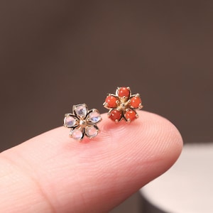 May include: Two small flower-shaped stud earrings made of gold. One earring features five translucent, iridescent white petals, while the other earring features five round, vibrant orange-red coral-like stones arranged in a floral pattern.
