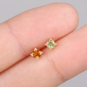 Op de afbeelding: Twee kleine oorstekers met een sterrenpatroon. Eén oorbel heeft een oranje edelsteen, de andere een groene edelsteen. Beide zijn gezet in goudkleurig metaal en worden op een vinger getoond.