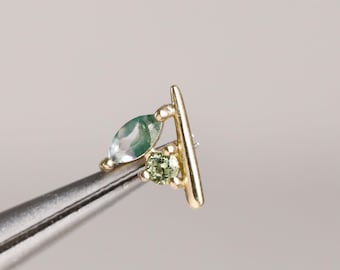 Könnte beinhalten: Ein goldener Ohrstecker mit einem vertikalen Stabdesign, akzentuiert durch einen grünen Stein im Marquise-Schliff und einen kleinen grünen Stein im Rundschliff. Das Schmuckstück wird von einer Metallpinzette gehalten und zeigt die feine Verarbeitung.