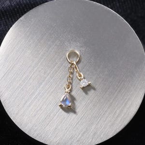 May include: A gold-colored charm with two dangling pendants. One pendant is a small, pear-shaped, iridescent stone, and the other is a small, triangular, clear stone. The charm is on a silver-colored, circular surface.
