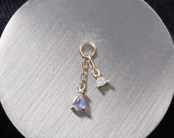 May include: A gold-colored charm with two dangling pendants. One pendant is a small, pear-shaped, iridescent stone, and the other is a small, triangular, clear stone. The charm is on a silver-colored, circular surface.