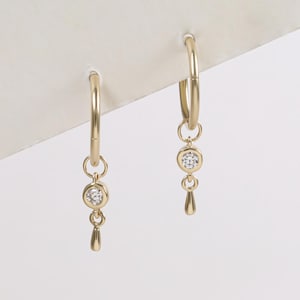 May include: A pair of gold hoop earrings. Each earring has a small, round, clear gemstone and a teardrop-shaped charm hanging from the hoop. The earrings are set against a white background.