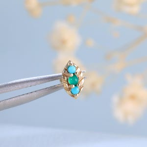 May include: A small gold stud earring featuring a marquise-shaped frame with a central round green gemstone flanked by two smaller round turquoise stones, held by metal tweezers to display the intricate metalwork and vibrant gemstone colors.