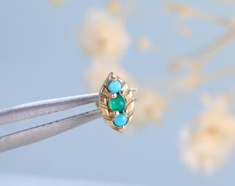 May include: A small gold stud earring featuring a marquise-shaped frame with a central round green gemstone flanked by two smaller round turquoise stones, held by metal tweezers to display the intricate metalwork and vibrant gemstone colors.