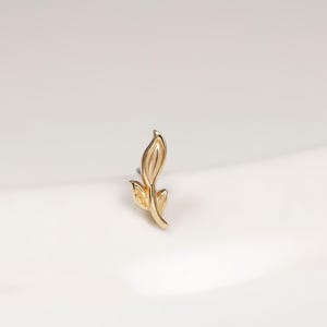 Könnte beinhalten: Ein kleiner goldener Ohrstecker in Form eines zarten Blattes mit polierter Oberfläche. Das Design zeigt eine mittige Blattader und eine spitz zulaufende Form, was für eine schlichte und klare Optik sorgt. Das Metall hat einen warmen Gelbton.