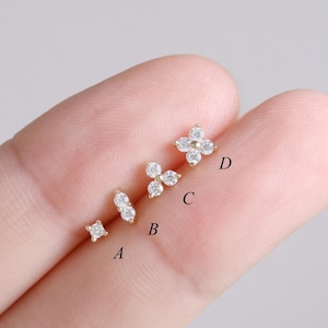 Puede incluir: Cuatro pequeños pendientes de oro con piedras preciosas claras y brillantes. Los pendientes están dispuestos en línea, con un cuadrado, un diseño de doble piedra, un grupo de tres piedras y una forma de flor de cuatro piedras. Fondo gris claro.