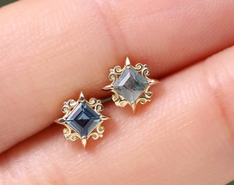 May include: Two stud earrings featuring square-cut blue gemstones set in gold metal frames. Each setting has a decorative, ornate border with four pointed star-like prongs extending from the corners, holding the central stone securely in place.
