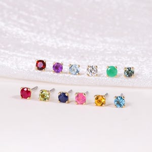 Puede incluir: Una colección de pendientes de botón con piedras preciosas de colores. Los pendientes están dispuestos en dos filas, mostrando una variedad de colores que incluyen rojo, morado, azul, verde y amarillo. Los pendientes están sobre un fondo blanco.