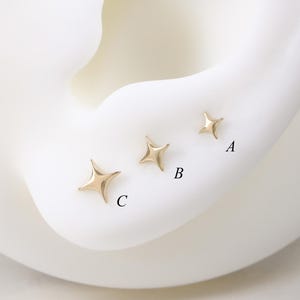 Puede incluir: Tres pendientes de botón dorados en forma de estrella de diferentes tamaños se muestran en un modelo de oreja blanco. Los pendientes están etiquetados A, B y C, siendo C el más grande y A el más pequeño. Los pendientes tienen un acabado brillante.