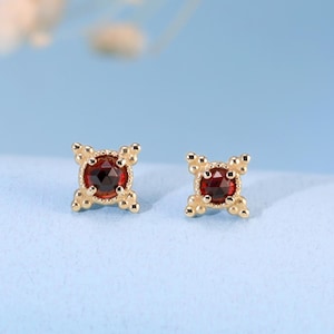 Può includere: Un paio di orecchini a lobo in oro con una pietra preziosa rotonda color rosso scuro incastonata in una montatura a forma di stella a quattro punte. Ogni punta della stella è decorata con tre piccole perline d'oro, creando un design simmetrico.