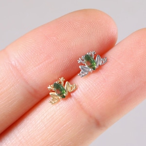 May include: Two small frog-shaped earrings. One is gold-toned, the other is silver-toned. Both have green gemstone eyes. The earrings are displayed on a light background.
