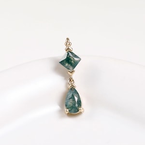 May include: A gold pendant featuring a kite-shaped moss agate stone positioned above a teardrop-shaped moss agate stone. The two green, translucent stones are set in gold metal settings and connected by a small gold link, creating a vertical drop.