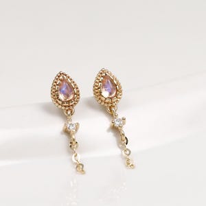 Peut inclure: Une paire de boucles d'oreilles pendantes en or avec une pierre irisée en forme de poire dans un cadre perlé. Chaque boucle possède une fine chaîne en or suspendue à la base, ornée d'une petite pierre ronde transparente à son extrémité.