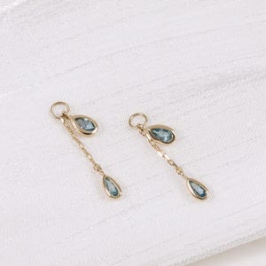 May include: Two gold-colored earrings with a delicate chain design. Each earring features a small, oval-shaped blue gemstone at the top and a teardrop-shaped blue gemstone at the bottom. The earrings are displayed on a white, textured surface.