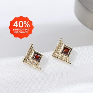 Peut inclure: Une paire de boucles d'oreilles dorées avec un motif géométrique. Chaque boucle d'oreille présente une pierre précieuse carrée, rouge foncé, sertie dans un cadre en forme de losange. Les boucles d'oreilles sont présentées sur une surface blanche, avec un autocollant rouge et blanc indiquant "40% LIMITED TIME DISCOUNT!"