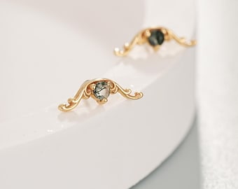 Könnte beinhalten: Zwei goldfarbene Ohrstecker mit einem herzförmigen grünen Edelstein in der Mitte. Die Ohrringe haben ein dekoratives, flügelartiges Design. Sie werden auf einer weißen, geschwungenen Oberfläche präsentiert.