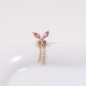 Könnte beinhalten: Goldener Ohrring mit Schmetterlingsdesign, besetzt mit rosa und lila Edelsteinen. Zwei goldene Ketten hängen von der Unterseite des Ohrrings. Der Ohrring befindet sich auf einer weißen Oberfläche.
