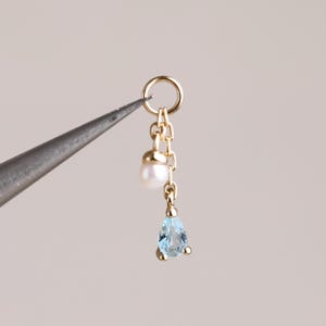 May include: A gold charm featuring a small white pearl and a light blue pear-shaped gemstone suspended from a delicate gold chain and a circular jump ring. The charm is held by a pair of metal tweezers, showcasing the fine metalwork and stones.