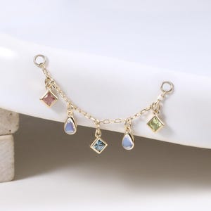 Puede incluir: Una delicada cadena de oro adornada con coloridos dijes de piedras preciosas. Los dijes incluyen piedras rosas, azules, verdes y transparentes en varias formas, incluyendo cuadrados y lágrimas. La cadena está diseñada para ser adjuntada a otras joyas.