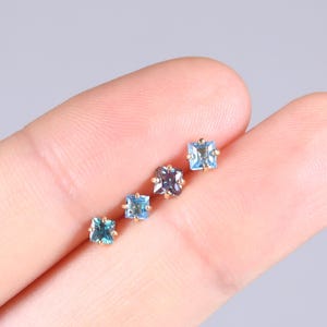 Puede incluir: Cuatro pendientes de botón con piedras preciosas cuadradas en diferentes tonos de azul y morado. Los pendientes están engastados en metal dorado. Las piedras están dispuestas en diagonal, mostrando sus variaciones de color.