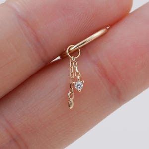 Könnte beinhalten: Ein goldener Creolen-Ohrring mit einem zarten, hängenden Kettenelement. Die Kette ist mit einem kleinen, klaren, rund geschliffenen Edelstein besetzt und endet in einem winzigen, glatten, tropfenförmigen Anhänger aus Gold.