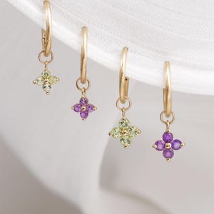 May include: Four gold hoop earrings, each with a small, four-petal flower charm. Two charms are purple, and two are green. The earrings hang against a white background.