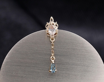 Puede incluir: Un pendiente de metal en tono dorado con un poste adornado de estilo filigrana engastado con una pequeña piedra ovalada transparente. Una delicada cadena dorada cuelga del poste y termina en un pequeño colgante de piedra azul en forma de gota.