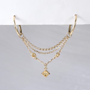May include: Gold-toned hoop earrings with a layered chain design. The chains are adorned with small, round, and square-cut yellow gemstones. The earrings hang from a white surface against a soft, blurred background.