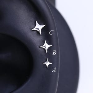 Puede incluir: Tres pendientes plateados en forma de estrella se muestran en un modelo de oreja negra. Los pendientes están dispuestos verticalmente, disminuyendo de tamaño de arriba a abajo, etiquetados A, B y C. Las estrellas tienen un diseño de cuatro puntas.