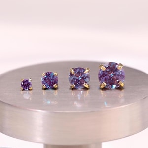 May include: Four round, faceted purple and blue color-changing gemstones set in gold-toned metal prongs. The stones are arranged in a row by size, from smallest to largest, each featuring a brilliant cut that reflects light with a vibrant shimmer.