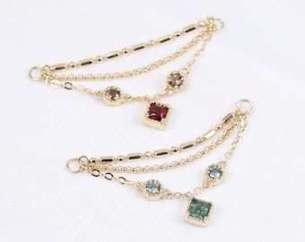 May include: Two gold chains with colorful gemstone pendants. The top chain has a red square gemstone and the bottom chain has a green square gemstone with two smaller blue round gemstones.