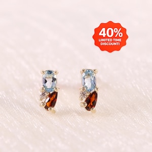 Könnte beinhalten: Ein Paar goldene Ohrstecker mit einem ovalen hellblauen Edelstein und einem birnenförmigen dunkelroten Edelstein, akzentuiert durch einen kleinen klaren Stein. Ein rotes rundes Abzeichen zeigt den Text 40% LIMITED TIME DISCOUNT!