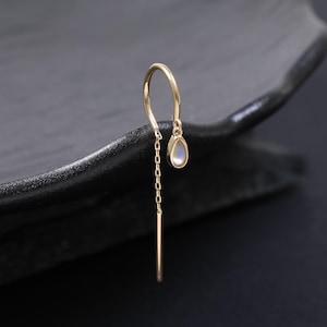 May include: A single gold earring with a small, teardrop-shaped gemstone. The earring features a delicate chain and a straight bar that hangs down. The earring is set against a dark, textured background.